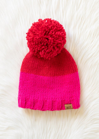 Two-tone red and pink knit beanie with a pom-pom on a white fur background