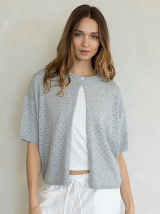 Woman wearing a gray knit top over a white shirt with white pants against a plain background