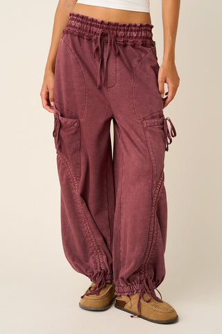 A person wearing loose, wide-leg cargo-style pants in a dusty burgundy or mauve tone, paired with tan lace-up shoes, against a plain light beige studio background.