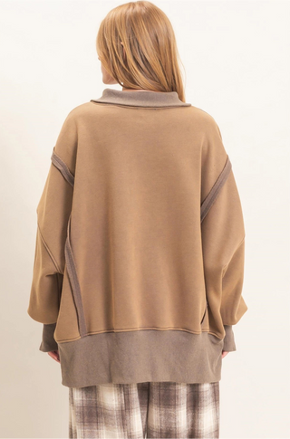 A model shown from the back wearing an oversized tan sweatshirt with contrasting brown ribbed trim and plaid pants, set against a plain off-white background.
