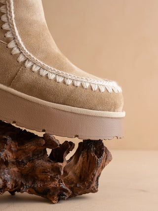 A close-up of a beige suede boot with white whipstitching and a chunky platform sole, resting on a dark brown piece of driftwood against a warm beige background.