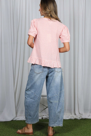 Person wearing a pink checkered top and blue jeans standing against a white curtain.
