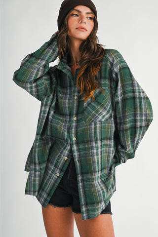 A woman wearing an oversized green plaid button-up shirt with black shorts and a dark beanie, photographed against a plain light gray studio background.