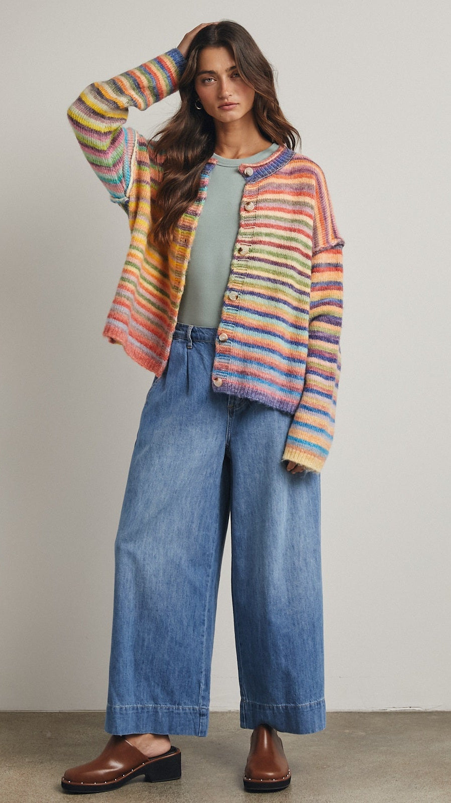 Woman wearing a colorful striped cardigan, green top, and blue jeans against a neutral background