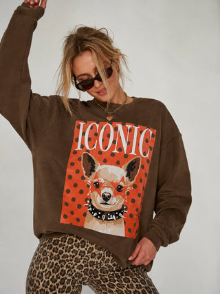 A woman wearing a brown sweatshirt with a large graphic print of a dog on an orange polka-dot background with the word 'ICONIC' in white, paired with leopard-print pants, against a plain light gray studio background.