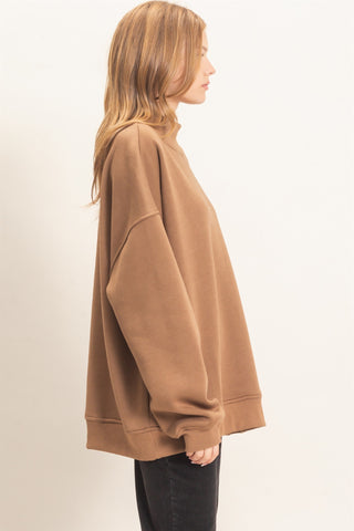 A woman shown in side profile wearing an oversized camel brown sweatshirt and black pants against a plain off-white studio background.