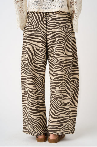 Wide-leg trousers with a brown and beige zebra print pattern, shown on a model from the waist down wearing brown sandals and a cream lace top.