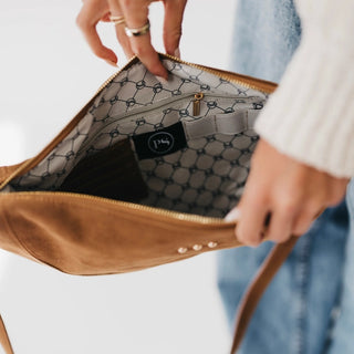 A close-up of a person holding open a tan suede shoulder bag to show the interior lining, which features a gray and black geometric pattern and a black brand patch reading 'PDA'.