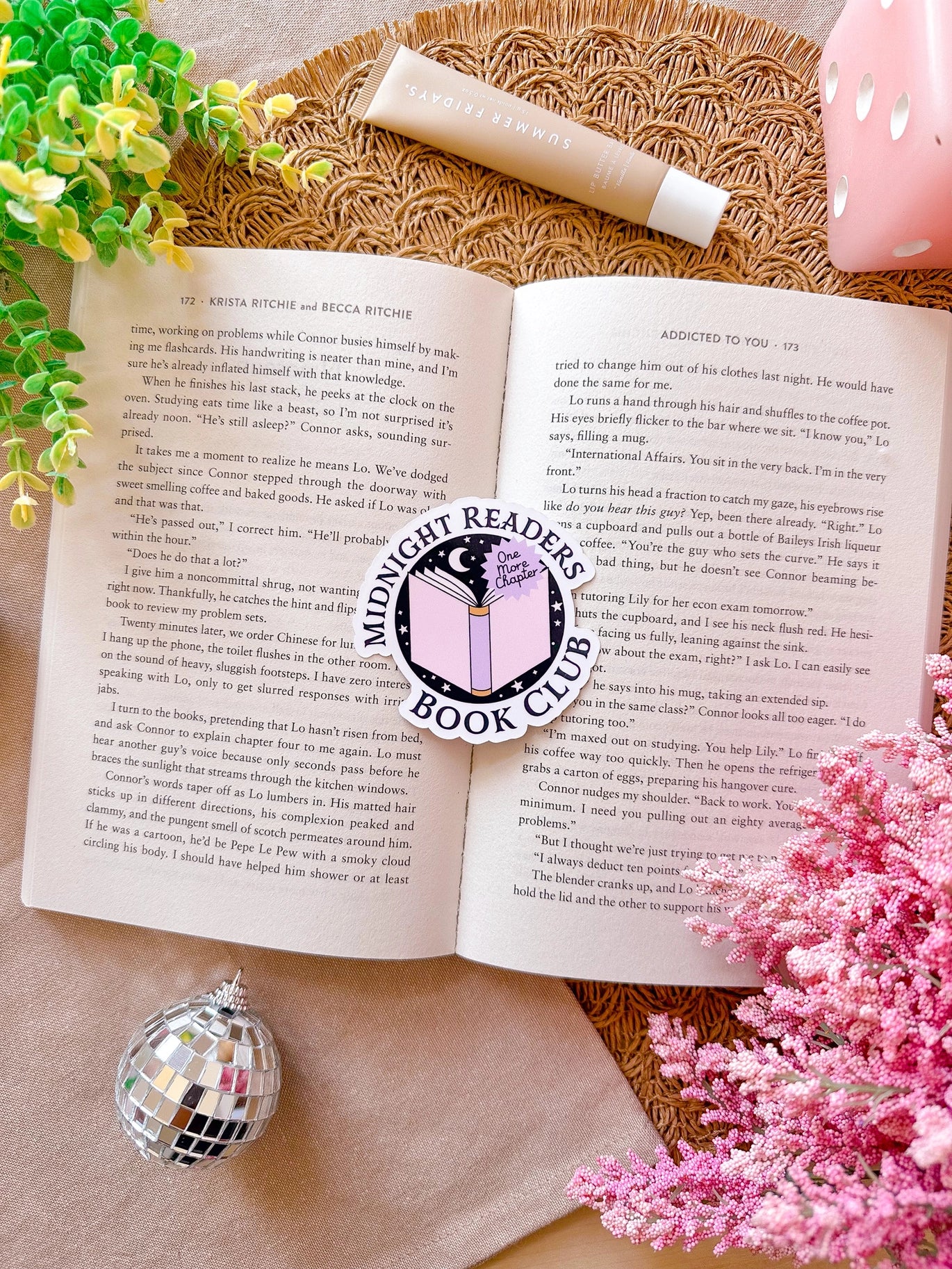 Open book with a decorative sticker on a textured surface surrounded by flowers and a disco ball.