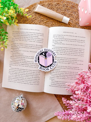 Open book with a decorative sticker on a textured surface surrounded by flowers and a disco ball.