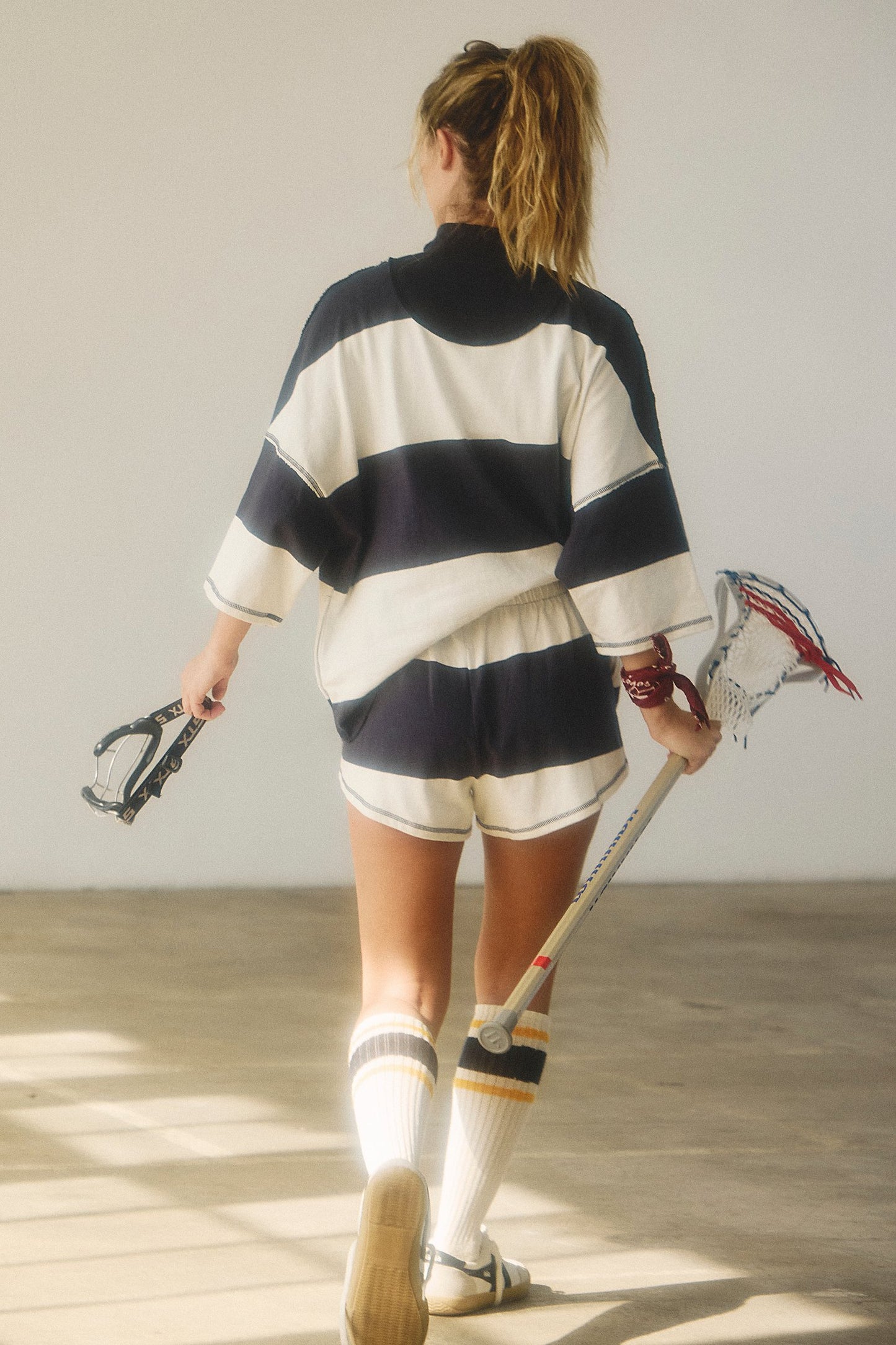 Person wearing a striped sweater and shorts holding lacrosse equipment indoors.