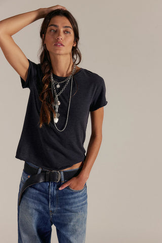 Person wearing a black t-shirt and blue jeans with layered necklaces on a neutral background