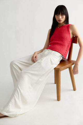 A model sits on a wooden chair wearing a bright red sleeveless knit top and wide-leg white trousers against a plain light gray studio background.