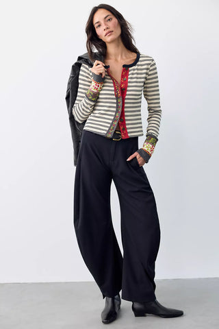 A model stands in a studio wearing a striped cropped cardigan over a red embroidered top, black wide-leg pants, and black boots, with a black jacket draped over one shoulder.