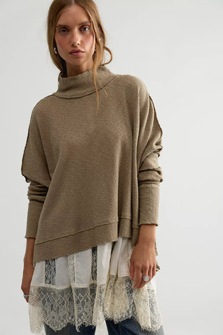 A woman wearing a taupe-brown ribbed knit turtleneck sweater with draped sleeves, layered over a white lace-trimmed dress, against a plain light gray background.