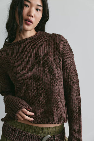 A woman wearing a dark brown ribbed knit cropped sweater with a wide boat neckline and long sleeves, paired with olive green bottoms, against a plain light gray background.