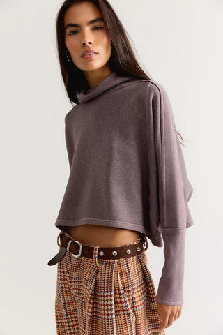 A woman wearing a cropped, long-sleeve cowl-neck sweatshirt in a muted mauve-purple color, paired with a brown studded belt and a brown, orange, and blue plaid skirt, against a plain light gray background.