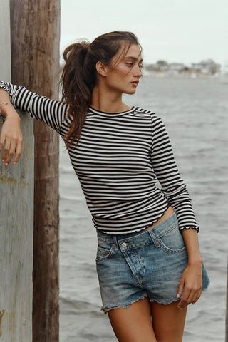 Woman in a striped shirt and denim shorts leaning against a wooden post by water.
