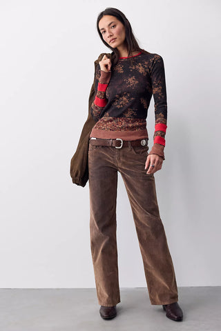 A woman modeling a brown corduroy wide-leg pantsuit with a matching brown jacket, a dark brown patterned long-sleeve top with red cuffs, and brown leather shoes, set against a plain light gray studio background.
