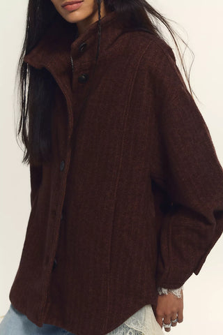 A close-up of a woman wearing a dark brown textured knit cardigan with a high collar and button front, paired with light blue jeans against a plain off-white background.