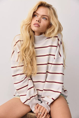 A blonde woman wearing a white long-sleeve mock neck sweatshirt with thin dark red horizontal stripes, sitting against a light gray background.