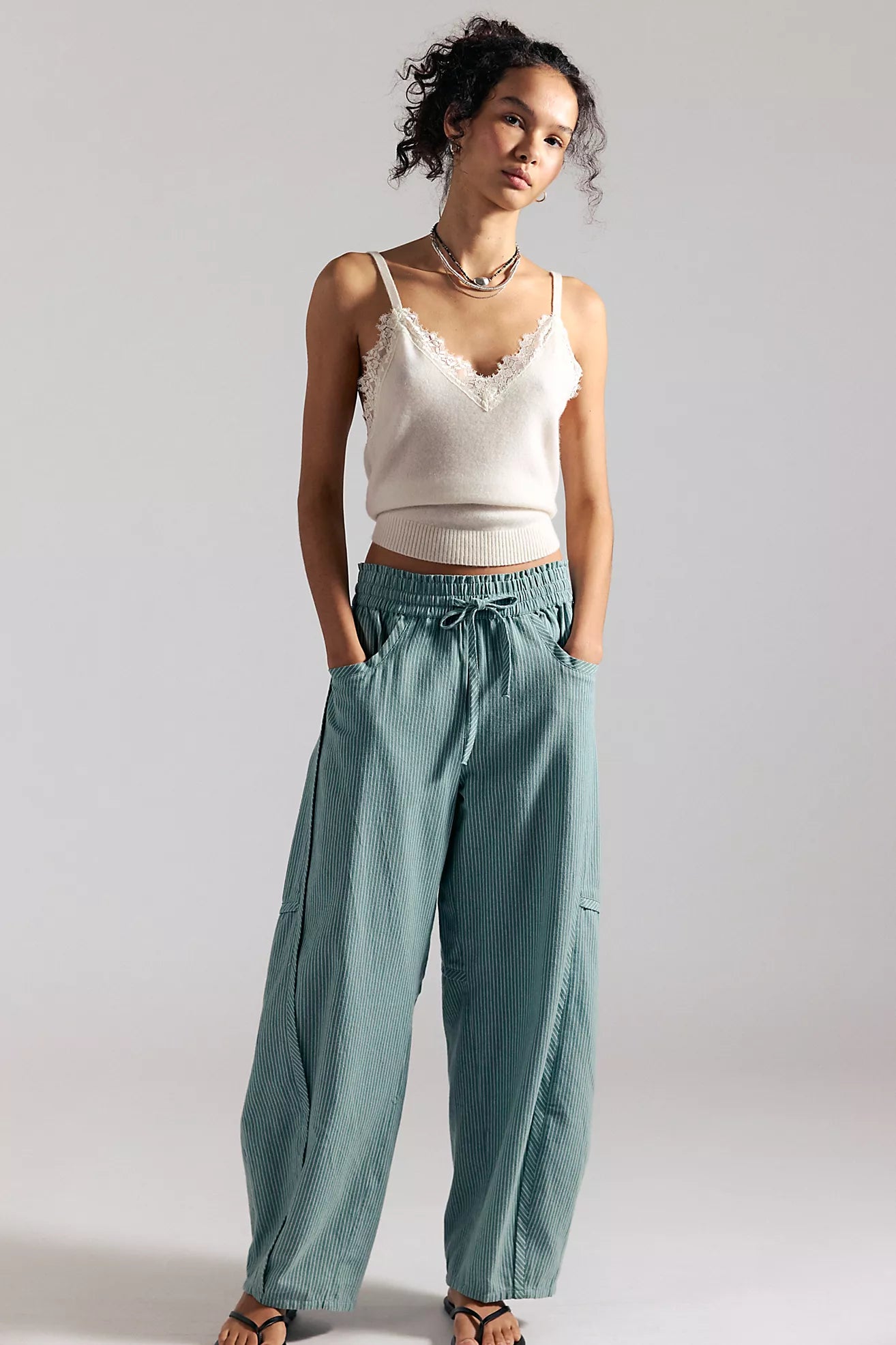 Woman wearing a white top and teal pants against a plain background