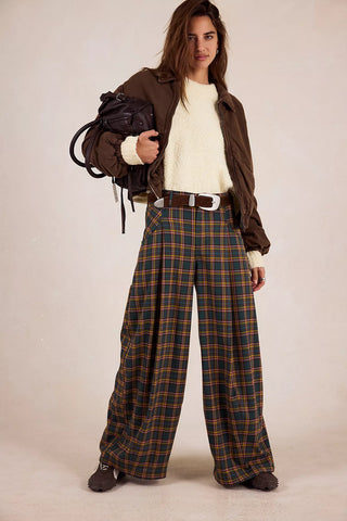 Person wearing a brown jacket, white shirt, and plaid pants holding a black bag against a plain background