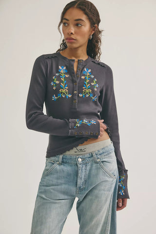 Woman wearing a dark gray long-sleeve top with floral embroidery and light blue jeans on a white background