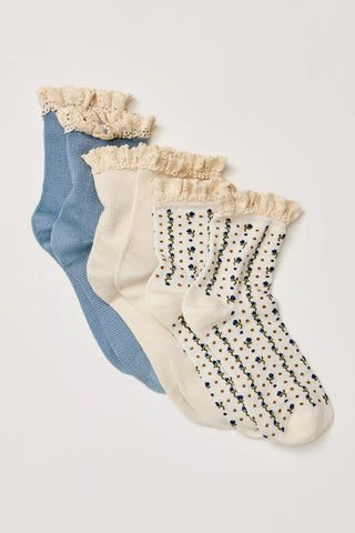 Three pairs of ankle socks arranged diagonally on a light beige background. The set includes a dusty blue pair, an off-white pair with a beige ruffle, and an off-white pair with a cream and blue floral pattern and a matching ruffle.