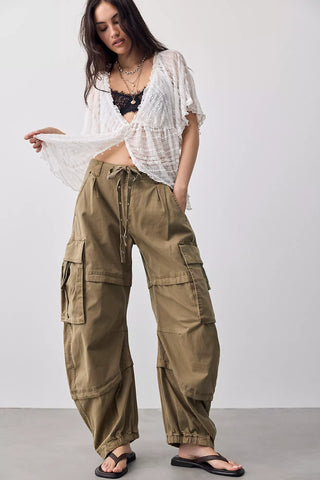 A woman modeling loose, wide-leg olive cargo pants with a drawstring waist, styled with a white sheer open-front top, black bralette, and black sandals against a plain light gray studio background.