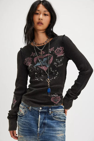 A model wearing a black ribbed long-sleeve top with a pink and white graphic design on the front, paired with blue jeans and layered necklaces, against a plain light gray background.