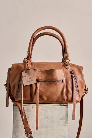A tan leather handbag with double top handles, front zip pockets, and a hanging tag reading 'WE THE PASS' is displayed on a textured pedestal against a plain beige background.
