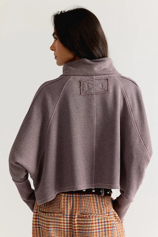 A model shown from the back wearing a cropped, long-sleeve high-neck pullover in a muted mauve-purple color, paired with a plaid skirt. The photo is taken against a clean, light off-white background.