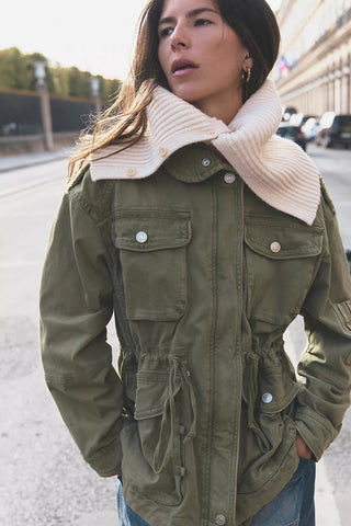 A woman wearing an olive green utility jacket with a large cream ribbed knit collar and a cream knit scarf, standing outdoors on a city street.