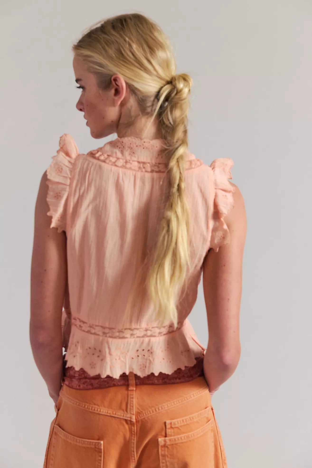 Woman wearing a light pink blouse with ruffled sleeves and orange pants against a plain background