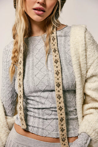 A close-up of a woman wearing a light gray cable-knit cropped top with a cream fuzzy cardigan and a patterned beige scarf, against a plain light background.