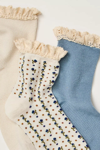 Three pairs of ribbed waffle-knit socks with cream ruffled trim, arranged diagonally on a white background. One pair is cream, one is light blue, and one features a cream base with vertical stripes of blue flowers and small brown dots.