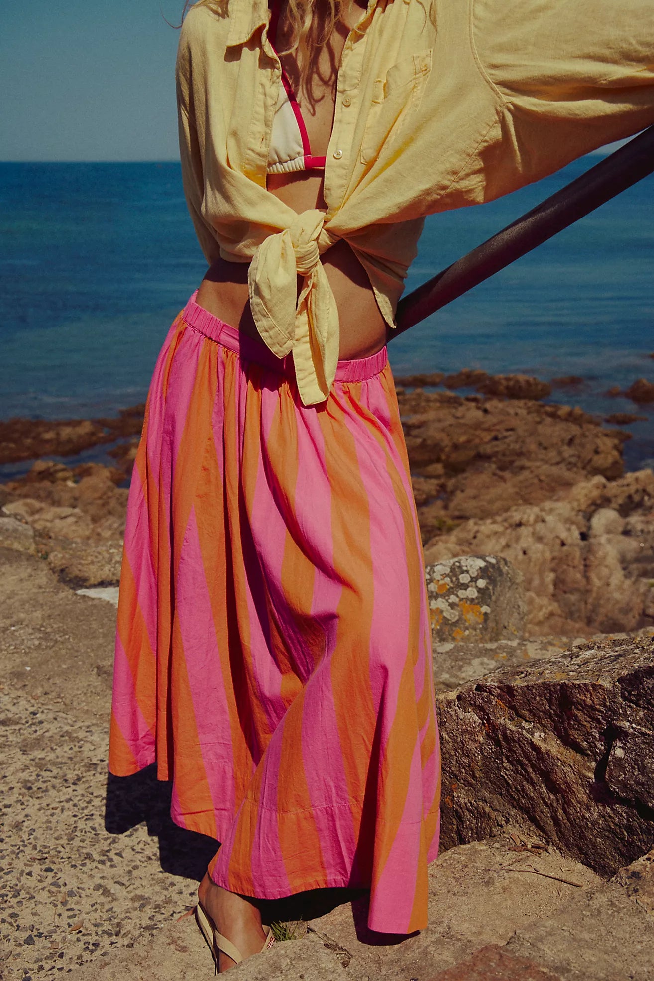 Person wearing a yellow jacket and colorful skirt standing on rocks by the ocean.