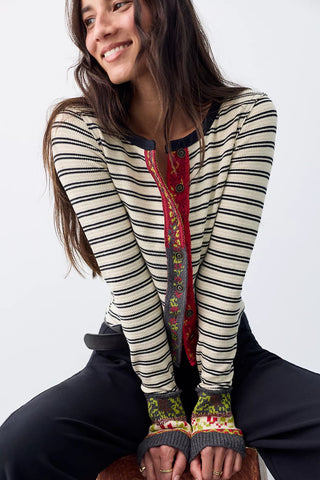 A woman wearing a cream and black horizontally striped long-sleeve top with a red and green paisley front panel, paired with black pants and knit fingerless gloves, sitting against a light gray background.