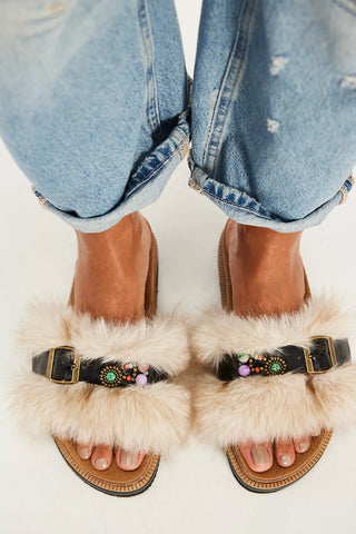 A top-down view of a person wearing light blue cuffed jeans and fluffy cream slide sandals with black straps, gold-tone buckles, and colorful bead embellishments on a white background.