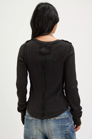 A woman viewed from the back wearing a black textured long-sleeve top with a small bow detail at the back neckline, paired with blue jeans against a plain white background.