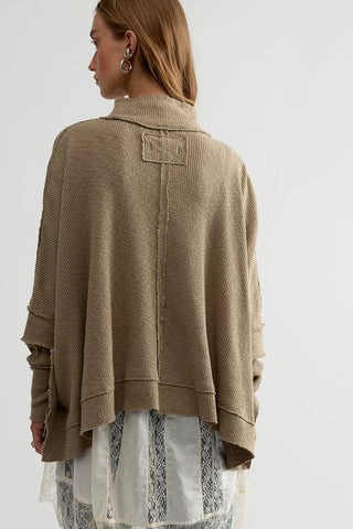A woman shown from the back wearing a loose, oversized ribbed knit sweater in a warm taupe-beige color, layered over a white patterned dress. The background is a clean, neutral off-white studio setting.