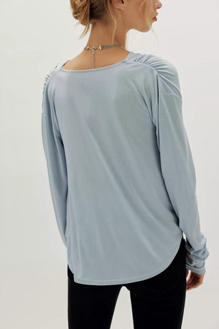 A woman shown from the back wearing a light blue long-sleeve top with gathered shoulder details and black pants, against a plain light background.