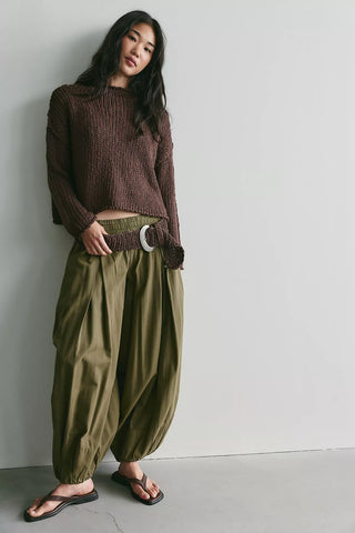 A woman stands against a plain light gray wall wearing a brown ribbed knit cropped sweater, olive green harem-style pants with a wide belt, and black flip-flop sandals.