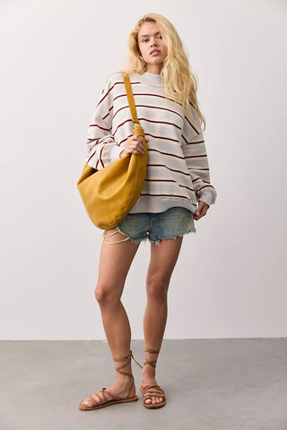 A woman stands in a studio wearing a cream and brown striped long-sleeve shirt, denim shorts, and brown strappy sandals, while carrying a large mustard yellow slouchy shoulder bag.