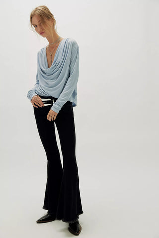 A woman modeling a light blue draped long-sleeve top with black flared pants and black shoes, standing against a plain light gray studio background.