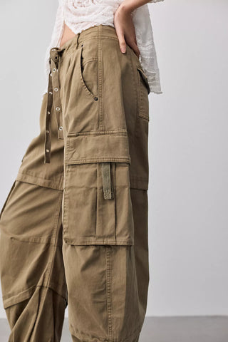 A close-up of a person wearing wide-leg olive cargo pants with multiple flap pockets, side drawstrings, and metal eyelet details, paired with a white lace-trim top against a light gray studio background.