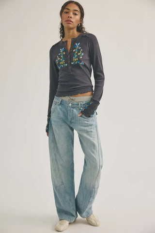 Woman wearing a dark gray long-sleeve top with floral embroidery and light blue jeans on a white background