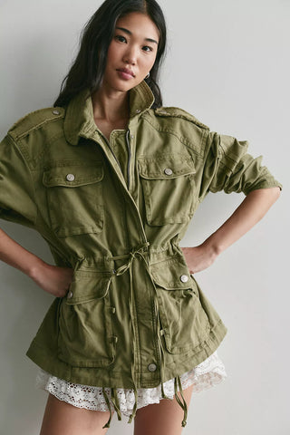 A woman modeling an olive green military-style utility jacket with a drawstring waist, front flap pockets, and silver snap buttons, paired with a white lace hem, against a light gray studio background.