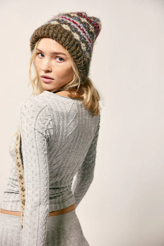 A woman wearing a gray cable-knit long-sleeve crop top and matching gray bottoms, paired with a brown knit beanie featuring red and white stripes, photographed against a plain light background.